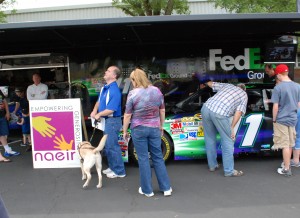 people standing outside with racecar in background and naeir sign next to it